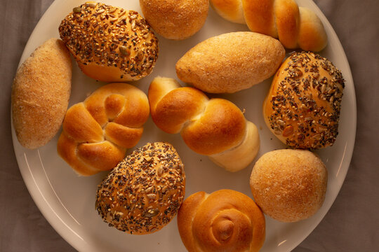 Overhead view of assorted baked bread rolls arranged on round white plate, including plain rolls, seeded rolls, and knotted rolls on neutral background.