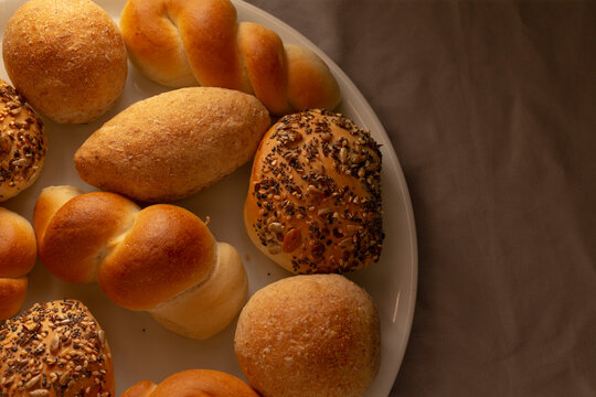 Close-up of assorted freshly baked bread rolls on white plate, with crusty textures and visible seeds, set against warm brown fabric background.