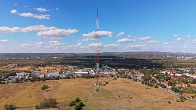 Aerial view of the historic Lakihegy radio mast rising above dry fields, industrial buildings and suburban homes in Szigetszentmikl&oacute;s, Hungary, beneath scattered clouds.