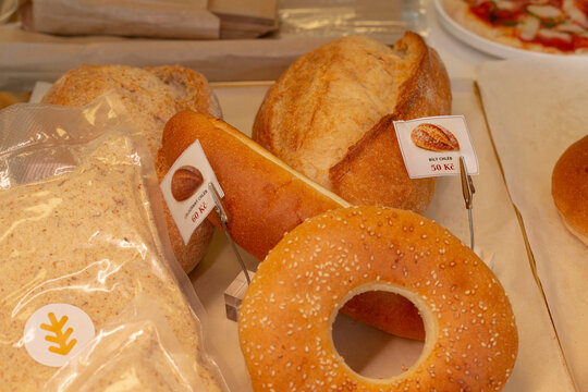 Assortment of baked goods including round seeded bread with hole, long roll, and crusty loaves displayed with price tags on counter.