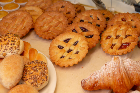 Close-up of assorted baked goods on countertop display, featuring lattice-top jam tarts, seeded rolls on plate, and powdered pastry edges.