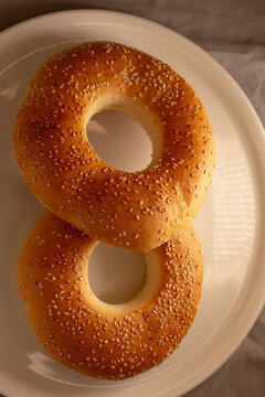 Overhead close-up showing two sesame seed bagels on white ceramic plate, highlighting golden crust texture, scattered seeds, and stacked portion of toasted bread.