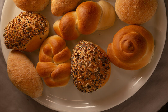 Overhead closeup of assorted freshly baked bread rolls and seeded buns on white ceramic plate, showing golden crusts and textured crumb for breakfast or snack.