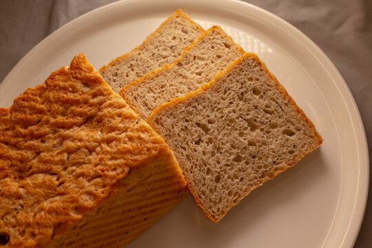 Close-up view of sliced whole grain bread on white plate, showing golden crust edges, porous crumb texture, and neat cut slices for breakfast or everyday meals.