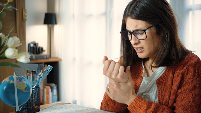 Young Latina student experiencing wrist tendinitis while studying at her desk. Concept of repetitive strain injury, wrist pain, study-related discomfort, prolonged writing