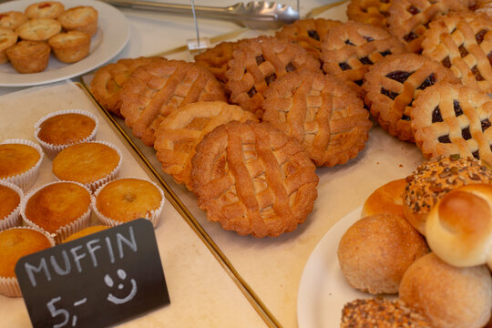 Close-up of assorted baked goods on display including multiple lattice-topped fruit tarts, paper-cup muffins, and various rolls arranged on trays and plates.