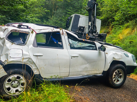 Heavily damaged white suv sitting by a forest road, being recovered by a crane after a serious vehicle accident