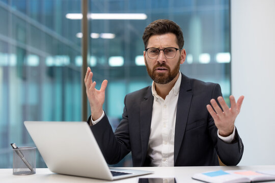 A young, disappointed businessman sits at his office desk, looking dejectedly at his laptop screen and spreading his hands in a gesture of frustration
