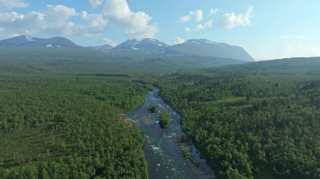 Aerial drone push in shot of the scenic Abiskojokk river flowing through a lush green valley in Sweden, surrounded by dense forest and distant snow-capped mountains along the Kungsleden trail.