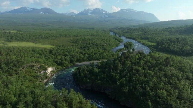 Aerial push in shot of the scenic Abiskojokk river winding through the lush green Abisko National Park with dense forest and mountains in the background, along the Kungsleden hiking trail in Sweden.
