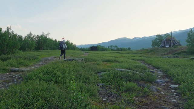 A solo hiker walks on the Kungsleden trail in Lapland, Sweden. Majestic mountains and lush vegetation surround a traditional Sami goahti hut and a trail marker, highlighting the peaceful adventure.