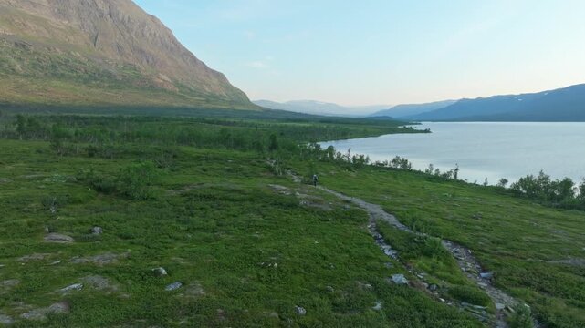 A push-in aerial drone shot shows a lone hiker on the Kungsleden trail in remote Lapland, Sweden. The stunning landscape features mountains, forests, and the pristine &Aacute;beskoj&aacute;vri lake.