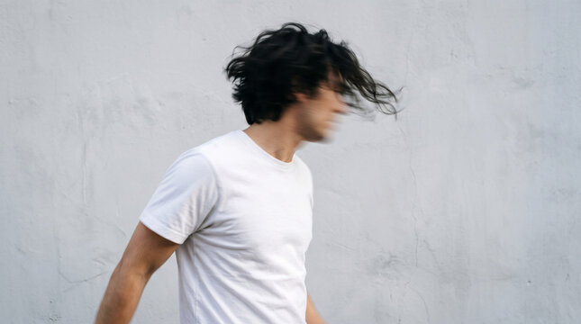 Man in white t-shirt with dark hair moving, creating a sense of motion and freedom against a textured grey wall, symbolizing energy and dynamism