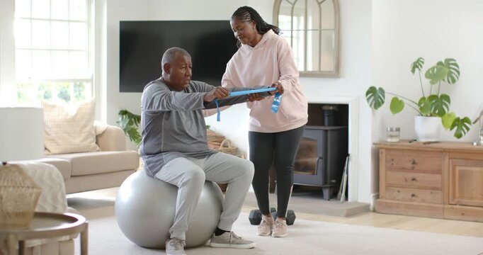 African American couple at home, woman taking band guiding man pressing on stability ball for rehab