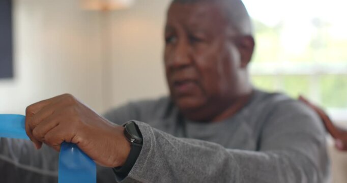 Senior African American man pulling blue band on couch, band taut, helper's hand bracing shoulder