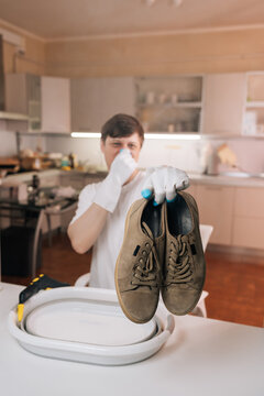 Vertical portrait of man wearing white gloves holding pair of stinky sneakers over laundry basin, covering nose due to foul odor preparing for cleaning in kitchen.