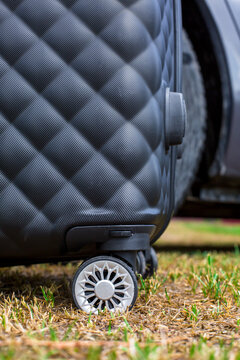 Black hard-shell suitcase with textured surface and wheel resting on grass near a vehicle, showcasing travel gear and outdoor setting