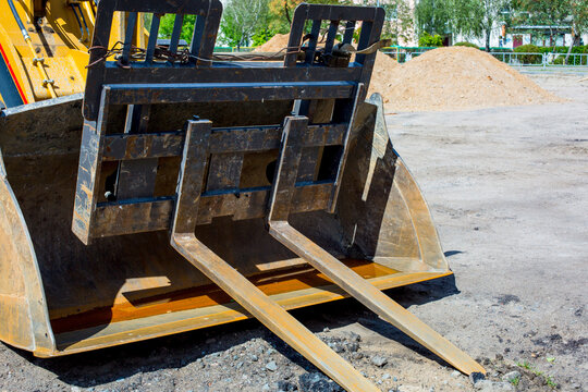 Heavy machinery bucket attachment with fork tines positioned on construction site, dirt piles and trees visible in the background under clear blue sky