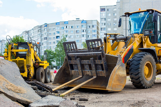 Heavy construction machinery including a yellow backhoe loader and a wheel loader working on urban infrastructure development near residential buildings