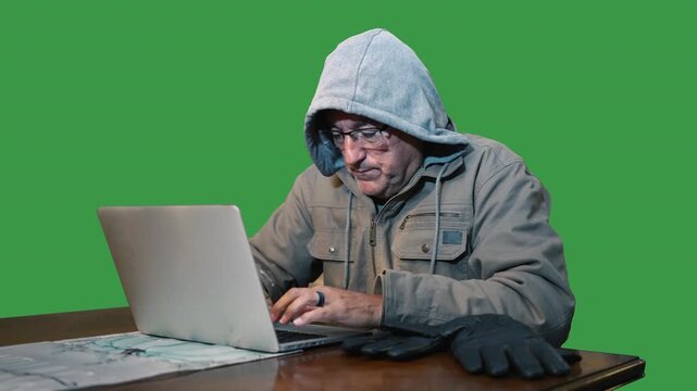 Man wearing a hooded sweatshirt and winter jacket types on a laptop while bundled up against the cold. Green screen background for easy compositing. 15446