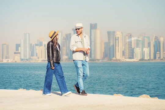 Doha Qatar tourist couple walking Corniche waterfront West Bay with skyline skyscrapers background happy tourists vacation lifestyle Middle East urban city exploration sunny winter fun day bonding