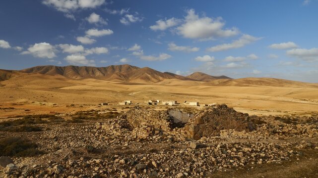 Island interior, barren landscape, reddish brown hills, ruins of buildings further away, ruin in foreground, blue sky, white clouds, Fuerteventura, Canary Islands, Spain