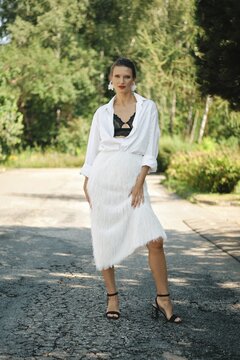 A fashion model poses confidently in a chic white fringed skirt and black top, complemented by a loose-fitting white shirt. The lush green background highlights her stylish presence on a sunny day
