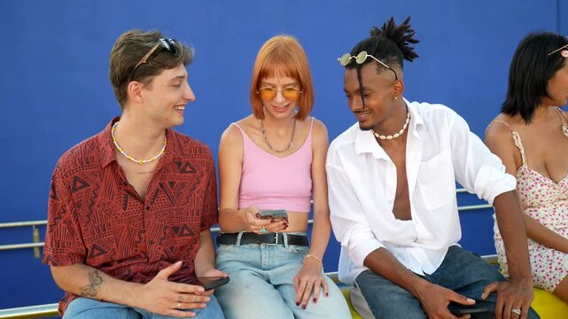 Group of stylish multicultural young friends sitting together and having fun while using their mobile phones, a girl showing something on her screen to the others against a blue background