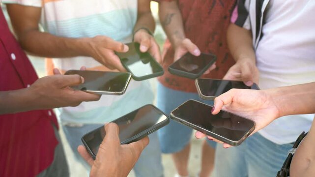 Diverse group of young friends standing together outdoors in a circle, deeply engaged and scrolling on their mobile smartphones, representing connection and modern social interaction