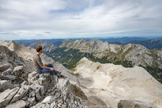 Hiker sitting at the summit of the Birkkarspitze, view of the Schlauchkar and Karwendeltal with the eastern Karwendelspitze, Hinterautal-Vomper chain, Karwendel, Tyrol, Austria