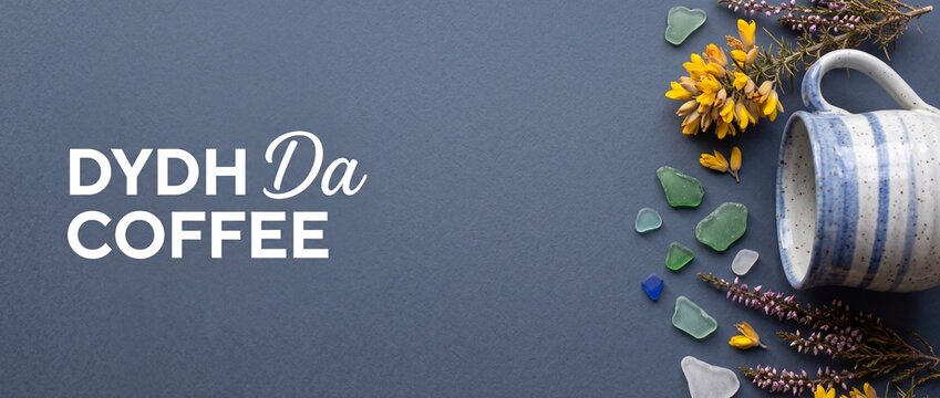 Cornish coffee mug flat lay banner background with sea glass, purple heather, yellow gorse flowers, and Celtic greeting text Dydh Da Coffee on textured blue paper for coastal cafe branding design.