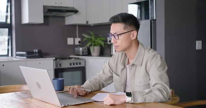 Asian man consulting laptop and writing notes at kitchen table for remote work with notebook