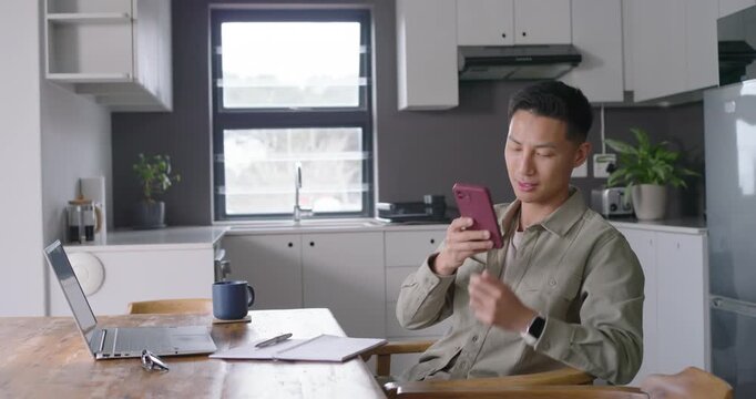 Asian man taking call on pink phone, jotting work notes, typing on laptop, copy space