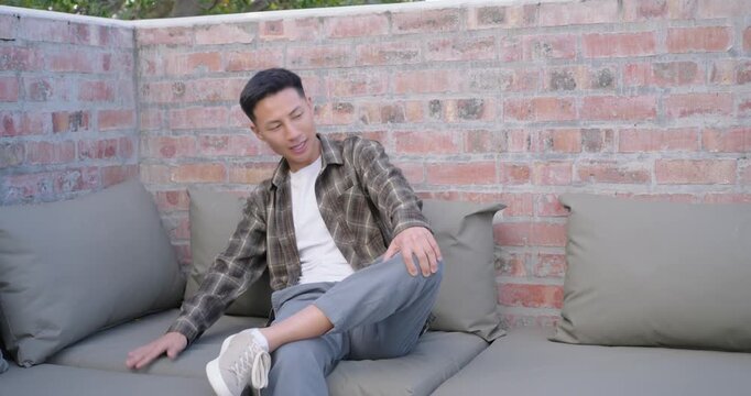 Mid-Adult Asian man settling on patio sofa, petting spaniel jumping with green tennis ball for play