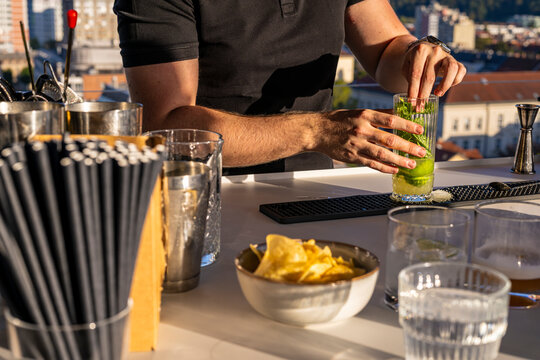 Bartender muddling fresh lime and mint in a tall glass while preparing a refreshing cocktail at a rooftop bar