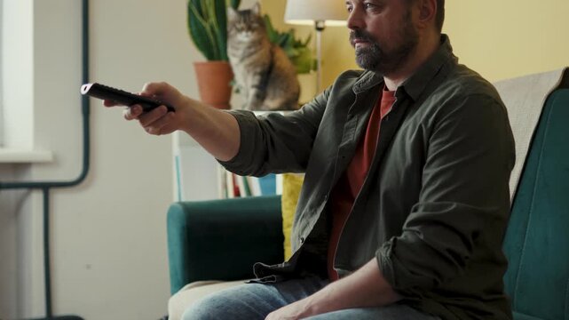 Middle-aged man switching channels with TV remote while sitting on couch with cat