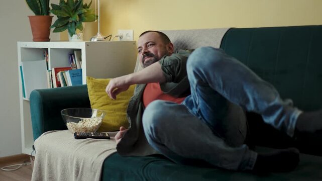 Lazy man lying down on couch reaching out for popcorn bowl on coffee table
