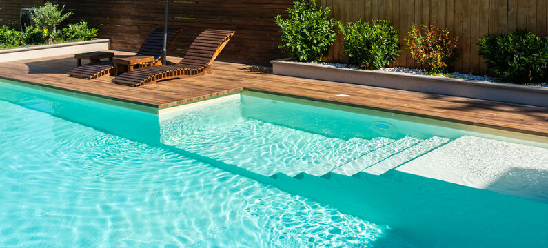 Modern swimming pool with turquoise water, wooden deck, and relaxing lounge chairs under sunny sky
