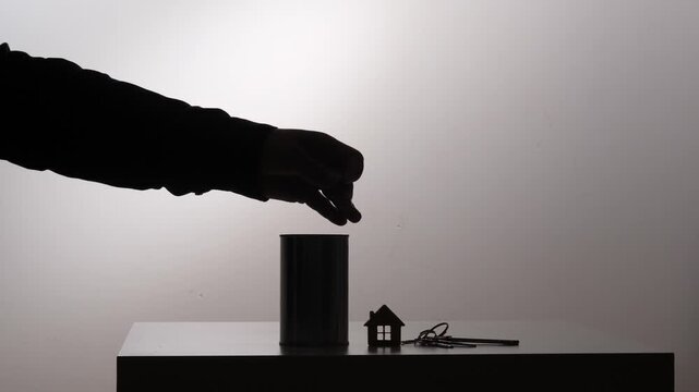 Hand silhouette dropping a coin into a piggy bank, symbolizing the act of saving money for real estate investment, home ownership, and future financial planning