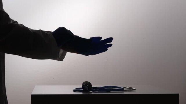 Doctor putting on a blue protective medical glove. Preparing for an examination or procedure. With a stethoscope resting on a surface below. Emphasizing healthcare. Safety. And medical professionalism