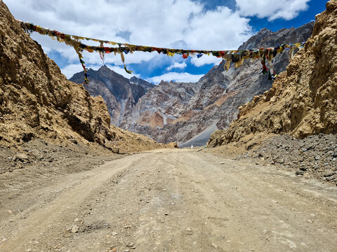 View of the rugged Penzi La road featuring colorful prayer flags strung across the path against a backdrop of towering mountain peaks under a cloudy sky in Zanskar, Ladakh, India.
