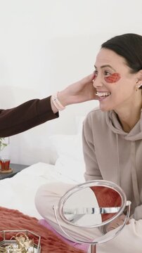 Vertical video: Smiling woman in tan hoodie having friend tuck hair on bed, with patches