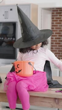 Vertical video: Reaching girl in witch hat cape sorting candies on kitchen island with pumpkin pail