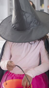 Vertical video: Smiling at cam, girl in witch hat reaching pumpkin pail to check candy in kitchen