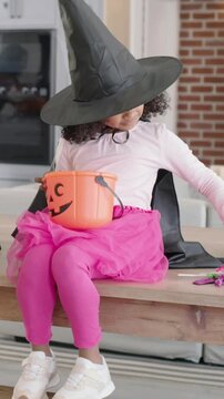 Vertical video: Spotting candies kid in witch hat rummaging pumpkin bucket in kitchen picking candy