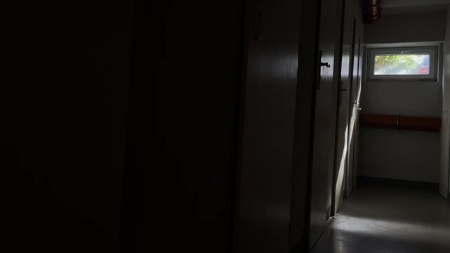 Dim interior corridor of school building during daylight, narrow hallway lined with closed wooden doors, small rectangular window at far end casting streak of sunlight across lin