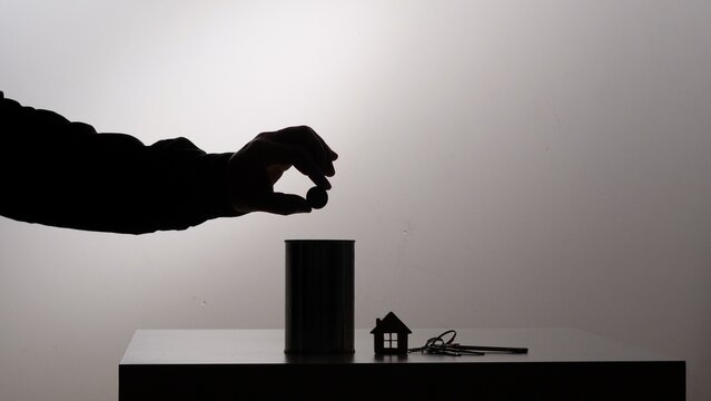 Hand dropping a coin into a savings can, symbolizing financial planning and investment towards the goal of purchasing a new house, with keys and a miniature home model resting on the surface