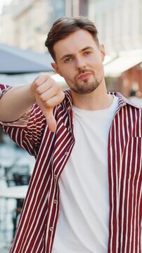 Dislike. Upset brunette bearded man showing thumbs down sign gesture, expressing discontent, disapproval, dissatisfied bad work, mistake. Displeased young guy standing in urban city street outdoors