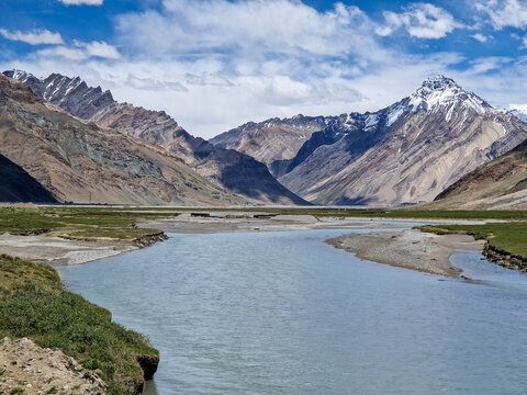 View of the Penzi La road winding through the dramatic Zanskar valley with snow-capped Himalayan peaks, a flowing river, and lush green meadows in Zanskar, Ladakh, India.