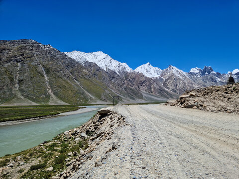 View of Penzi La road running alongside a river with snow-capped mountains in the background under a clear blue sky in Zanskar, Ladakh, India.
